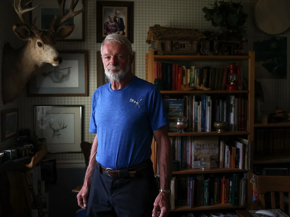 A portrait of Frank de Boon, a retired conservation officer, standing in his home office. He's wearing a blue t shirt, there's a bookshelf and photos on the wall behind him and a deer bust on the wall beside him.