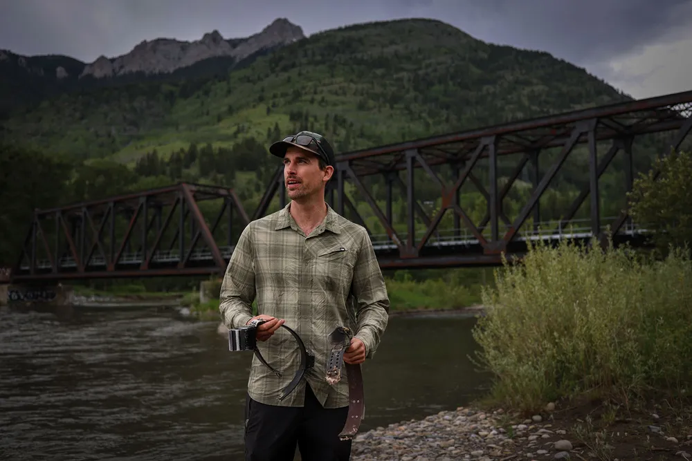 A photo of wildlife scientist Clayton Lamb holding damaged GPS collars on the edge of the Elk River near a rail bridge with mountains behind