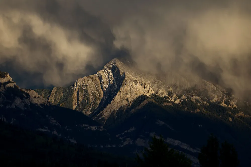Sunlight casts a glow against a rocky mountain peak shrouded in clouds.