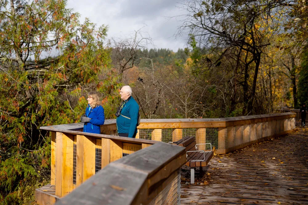 A photo of Jim Atkinson and his wife Judy Taylor-Atkinson on a boardwalk, covered with fallen leaves. They both have binoculars.