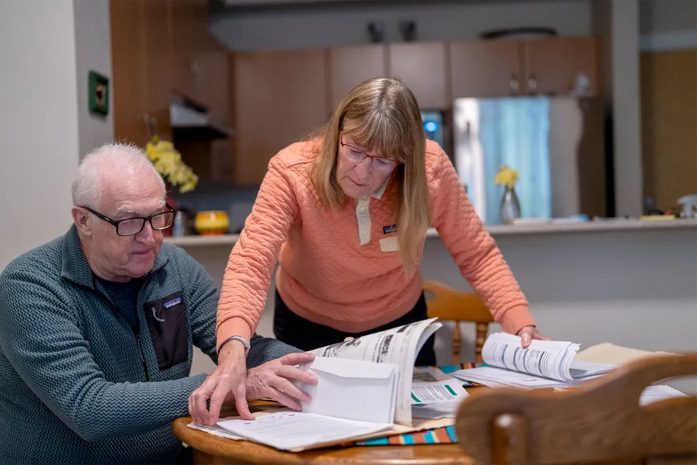 A photo of Jim Atkinson and Judy Taylor-Atkinson at their kitchen table, which is covered in documents. Judy is leaning in front of Jim flipping through a file.