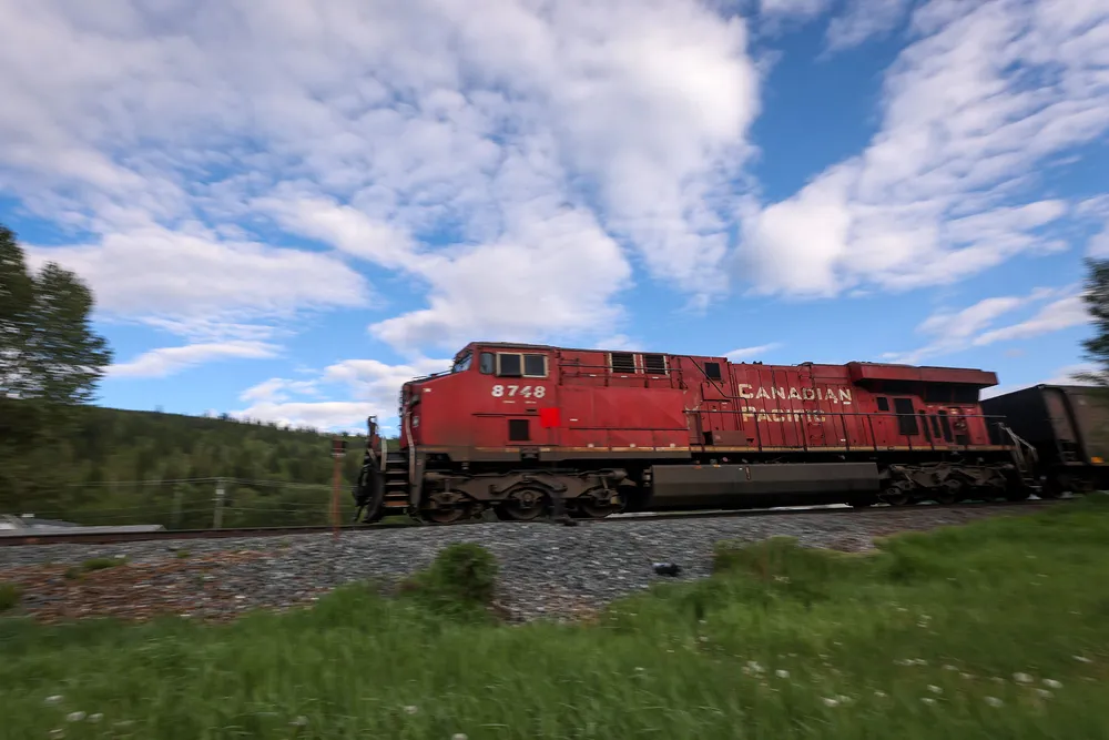 A red train locomotive chugging through Fernie BC with blue cloudy skies in the background. The photo is blurred by the train's movement. There are growing concerns about train wildlife collisions.
